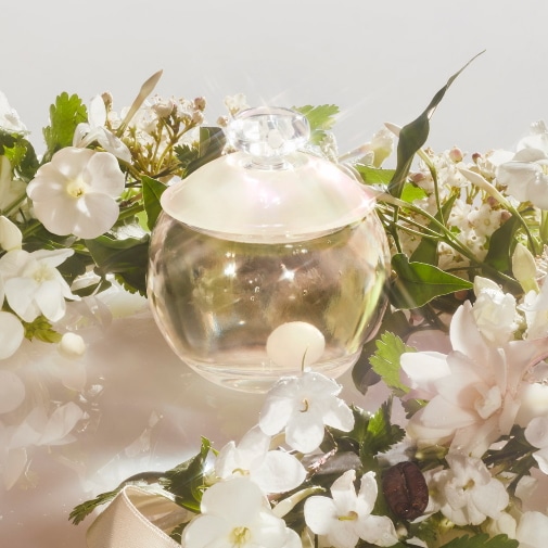 Okrągły flakon perfum z perłową nakrętką otoczony białymi kwiatami i zielonymi liśćmi tworzy romantyczną kompozycję.