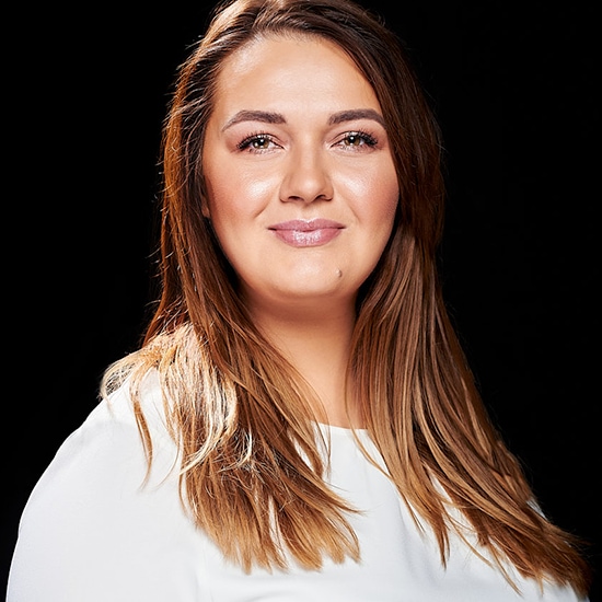 Portret kobiety z długimi, brązowymi włosami i w białej bluzce, na czarnym tle.