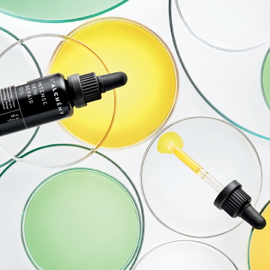 Butelka D'ALCHÉMY Intense Skin Repair Oil z pipetą na tle szalek Petriego wypełnionych żółtym i zielonym płynem.
