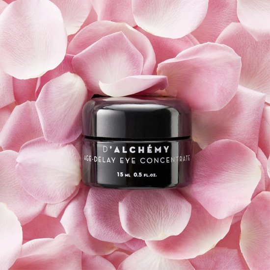 Czarny słoiczek D'Alchemy Age-Delay Eye Concentrate o pojemności 15 ml otoczony jest delikatnymi, różowymi płatkami kwiatów, tworząc elegancką kompozycję.