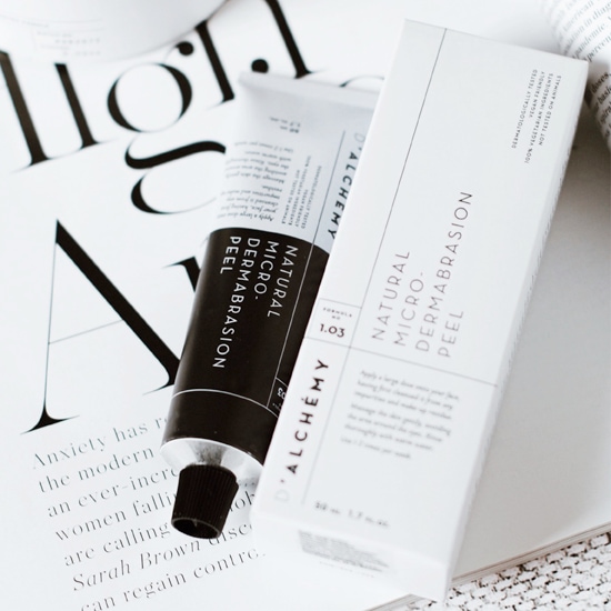 Peeling D'ALCHEMY Natural Micro Dermabrasion w białym opakowaniu i tubce, obok gazety, oferuje naturalny mikropeeling.