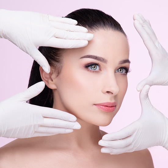 Na różowym tle twarz kobiety otoczona dłońmi w białych rękawiczkach, co sugeruje zabieg kosmetyczny lub dermatologiczny.