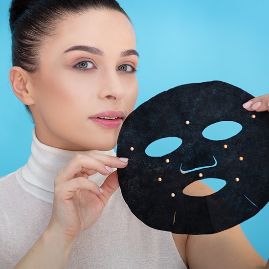 Kobieta trzyma w dłoni czarną maskę na twarz z otworami na oczy i usta, ozdobioną złotymi kropkami, na tle błękitnego tła.