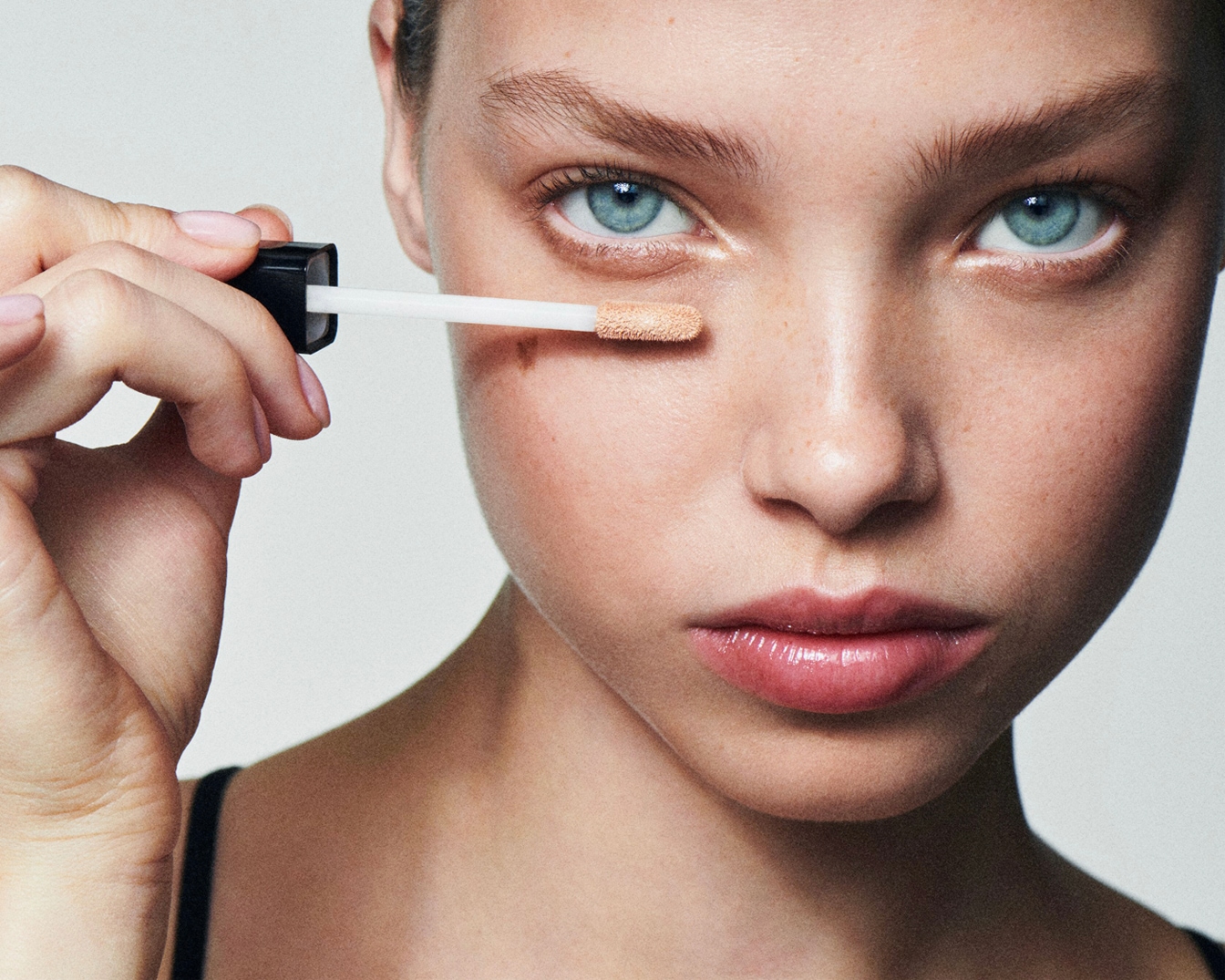 Nahaufnahme eines jungen, hellhäutigen Models mit strahlend blauen Augen, das mit einem Applikator Concealer unter dem Auge aufträgt.