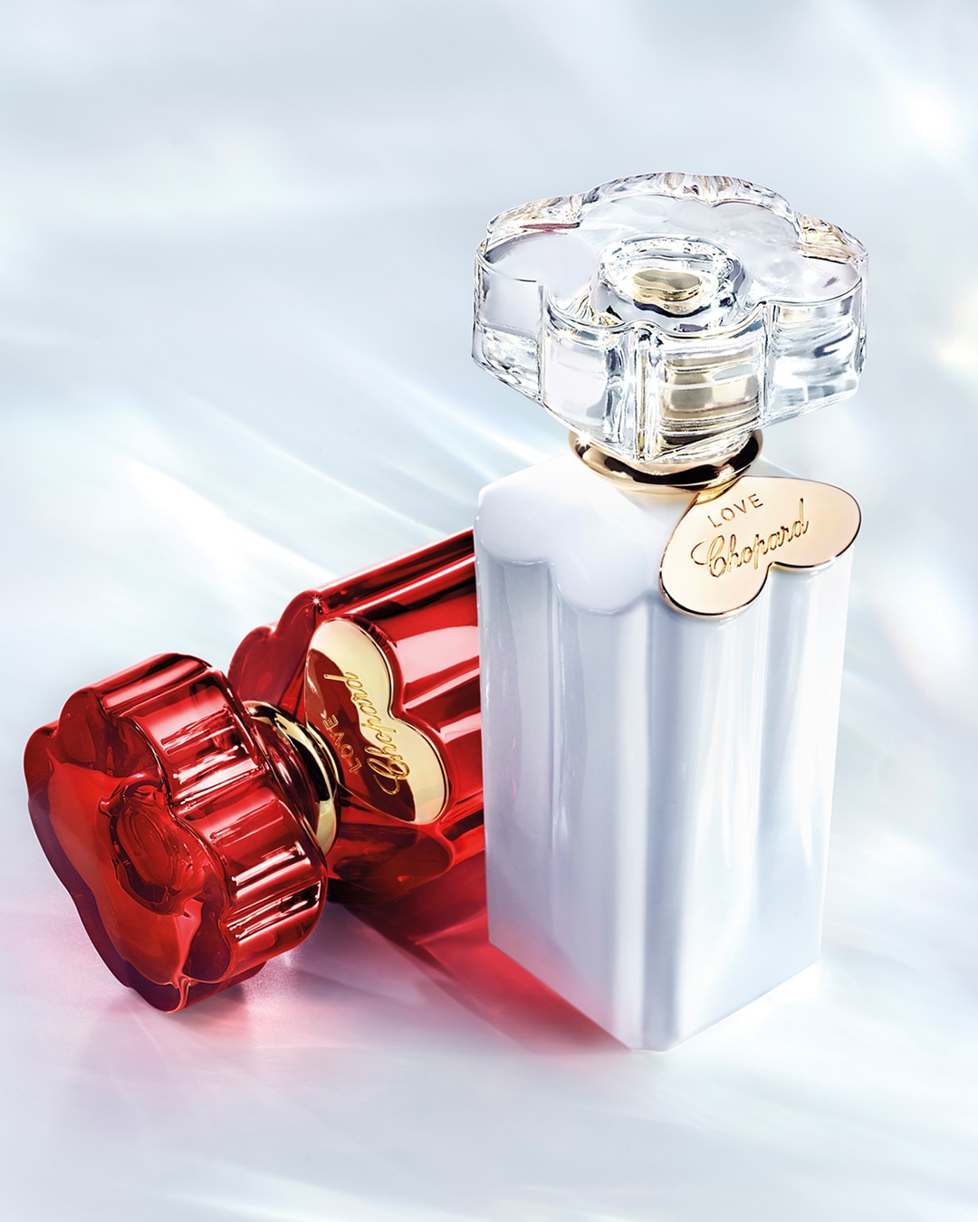 Dwa flakony perfum Love Chopard: biały i czerwony, oba z kwiatowymi korkami i złotymi zawieszkami z napisem Love Chopard.