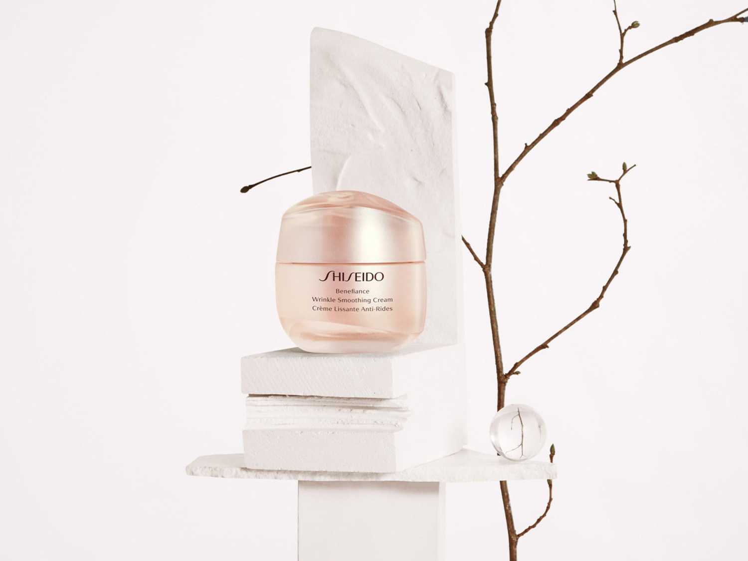 De Shiseido Benefiance Wrinkle Smoothing Cream in een zachtroze potje, gepresenteerd op witte stenen met een decoratieve tak, belooft een gladde, rimpelvrije huid.