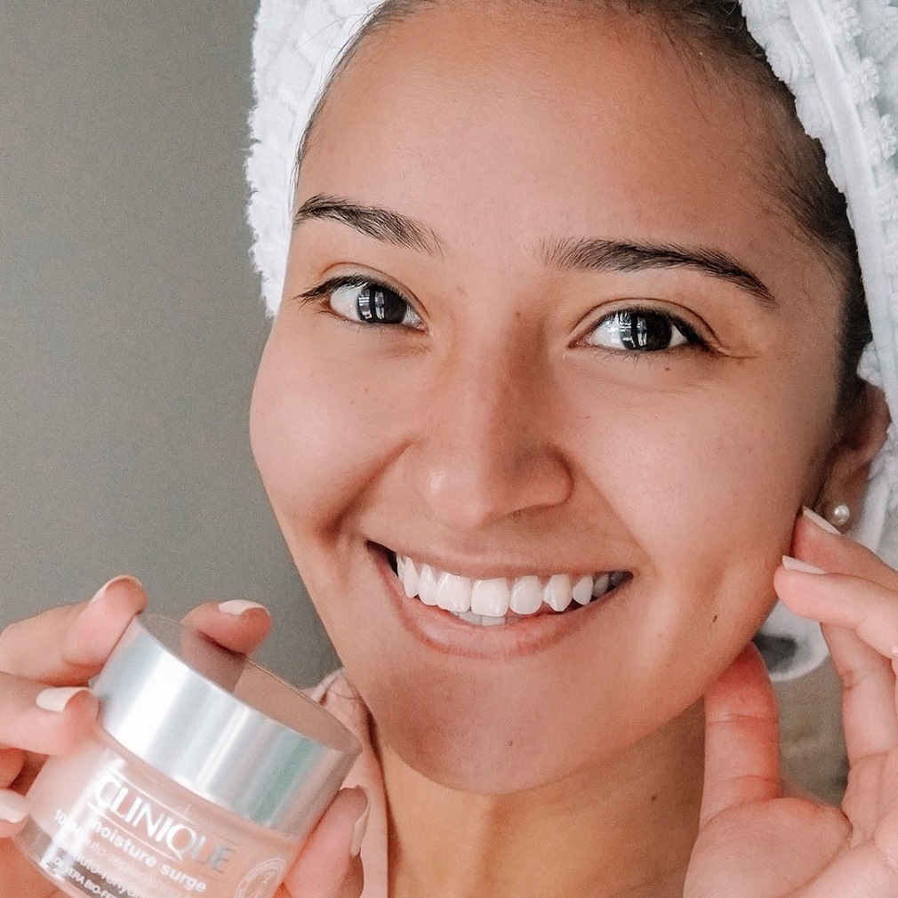 Une femme souriante, une serviette sur la tête, tient un pot de crème hydratante Clinique, prête à dorloter sa peau.