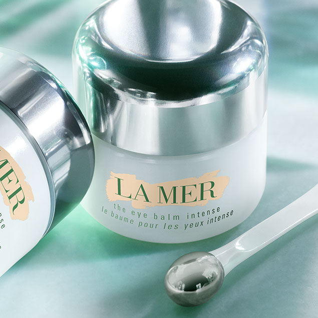 Voici le baume pour les yeux intense de La Mer, présenté dans un pot blanc élégant avec un couvercle argenté réfléchissant, accompagné d'un applicateur argenté.