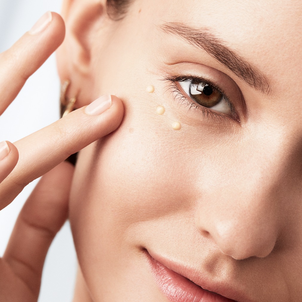 Gros plan sur une femme appliquant délicatement une crème beige sous son œil avec son doigt, pour une peau éclatante et hydratée.