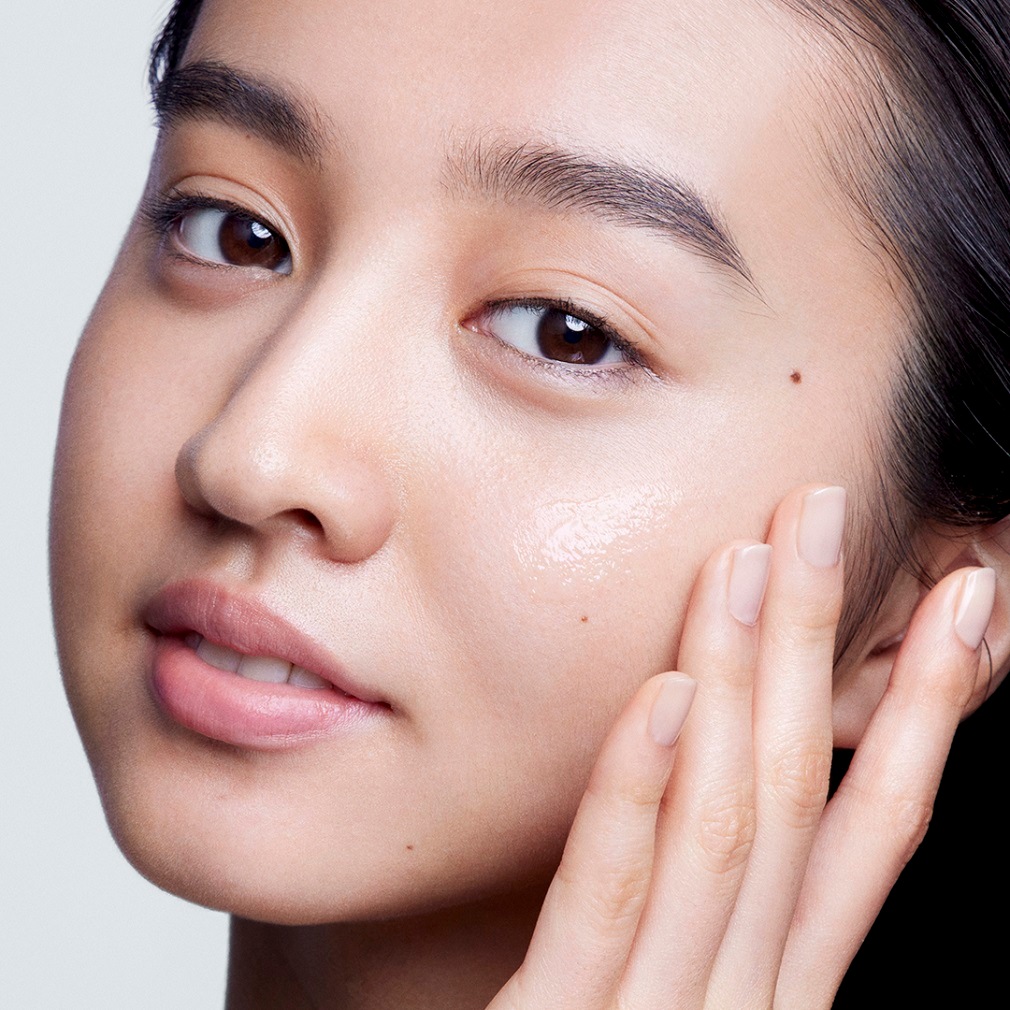 Une femme asiatique applique un sérum transparent sur sa joue, sa peau est claire et ses ongles sont vernis d'une couleur nude.