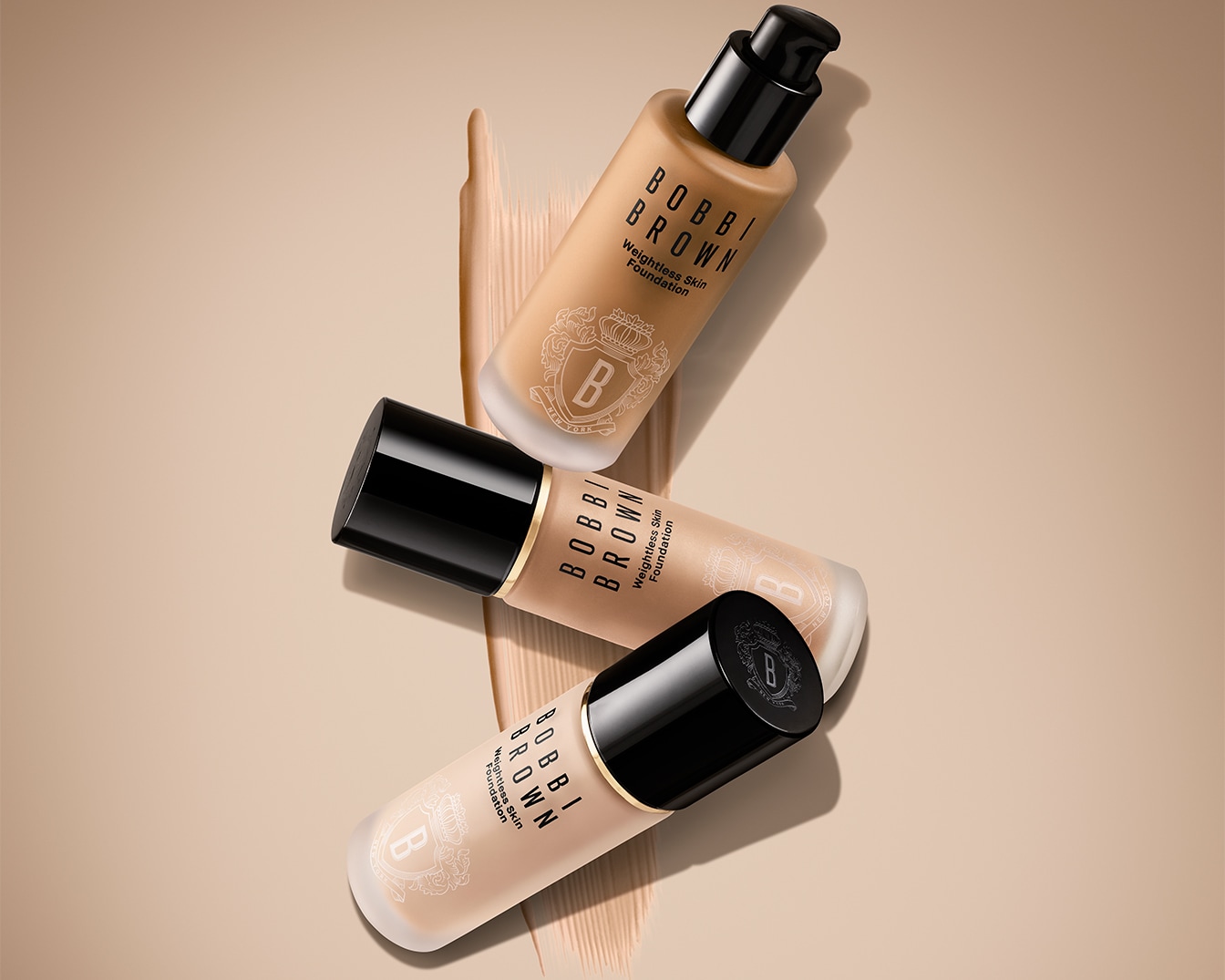 Három Bobbi Brown Weightless Skin Foundation üvegcséje látható, bézs színű alapon, fekete kupakkal és a márka logójával.
