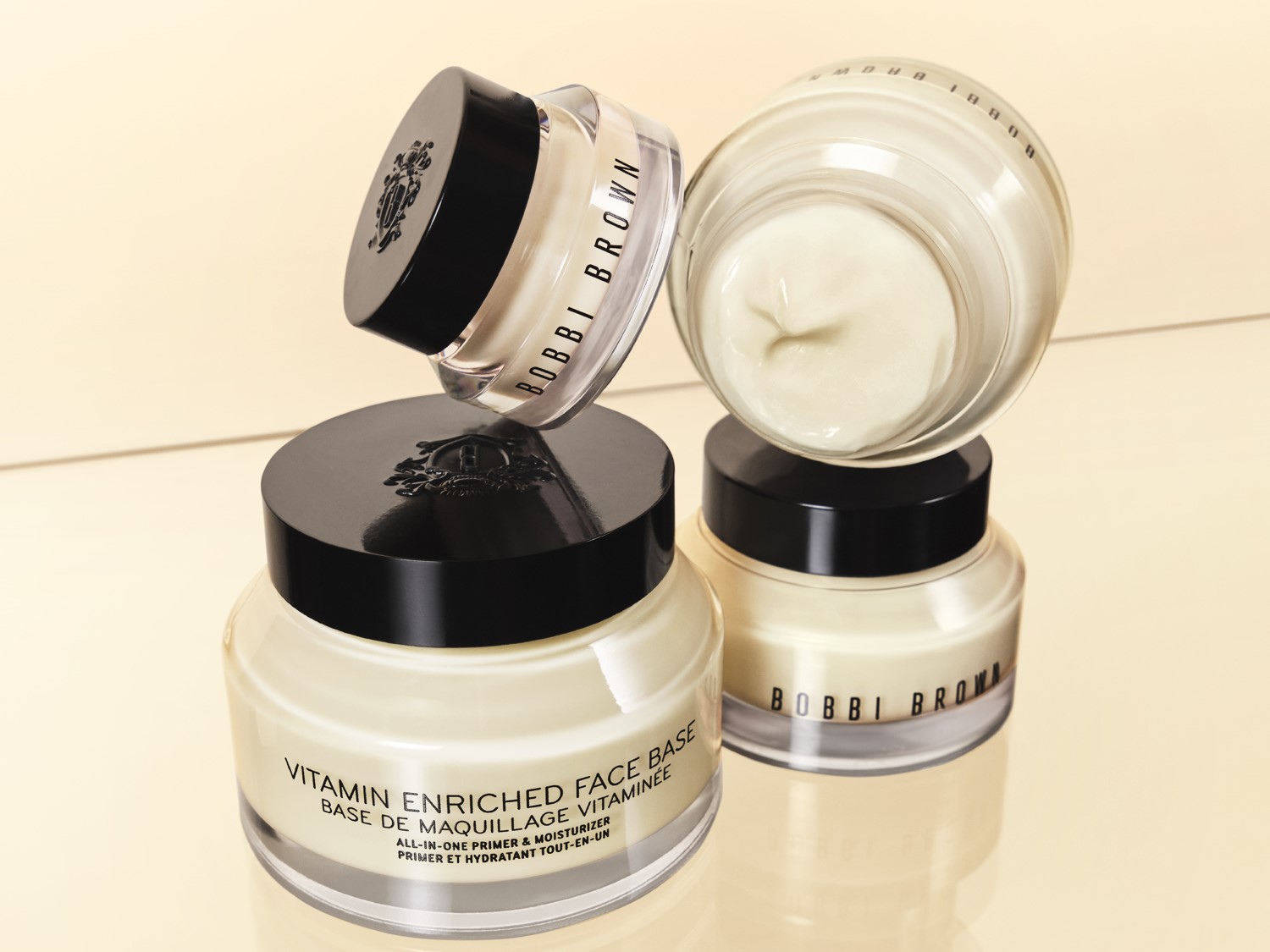 Kremowa baza pod makijaż Bobbi Brown Vitamin Enriched Face Base w przezroczystym słoiczku z czarną zakrętką, obok której znajduje się otwarty słoiczek z widoczną zawartością.