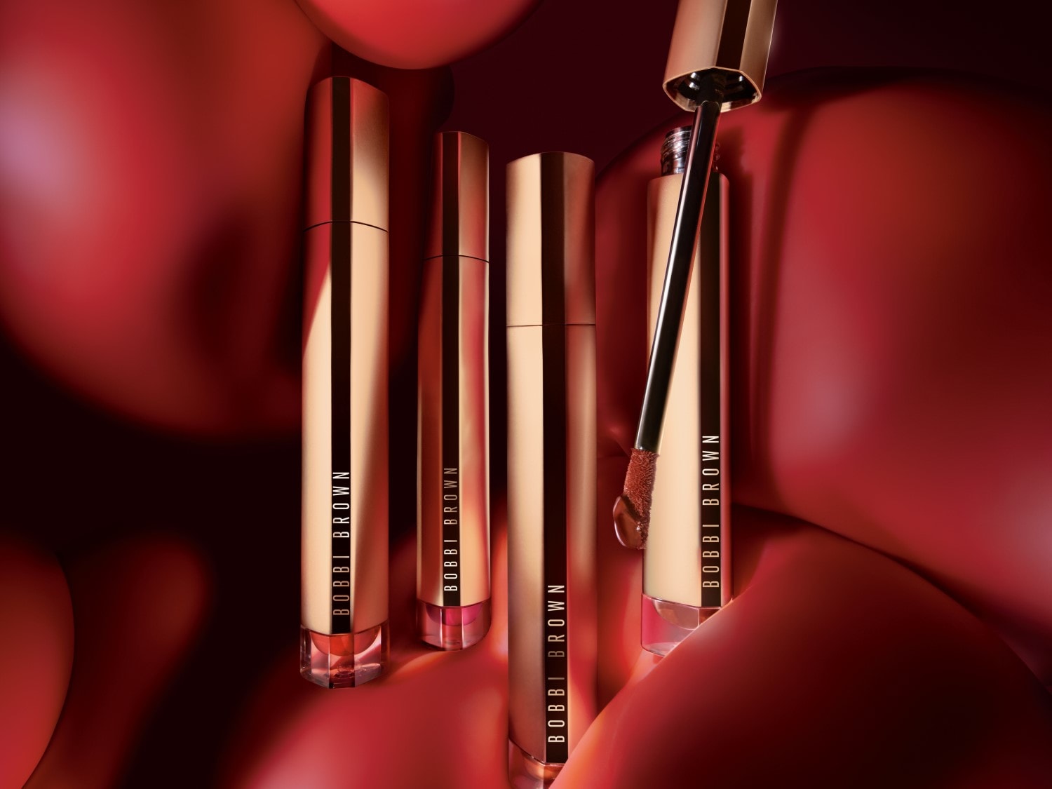 Cztery błyszczyki do ust Bobbi Brown w złotych opakowaniach na czerwonym tle, jeden z aplikatorem wyciągniętym, ukazującym odcień.