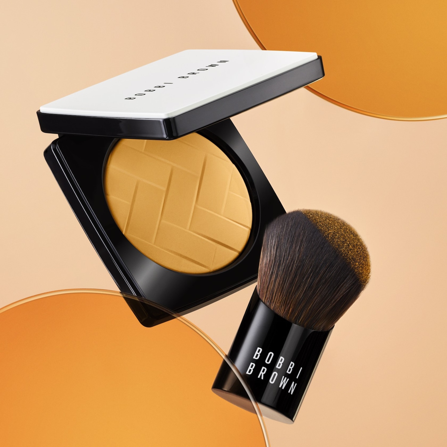 A Bobbi Brown púderes alapozója fekete tokban, fehér fedéllel, egy hozzáillő ecsettel lebeg egy narancssárga háttér előtt.