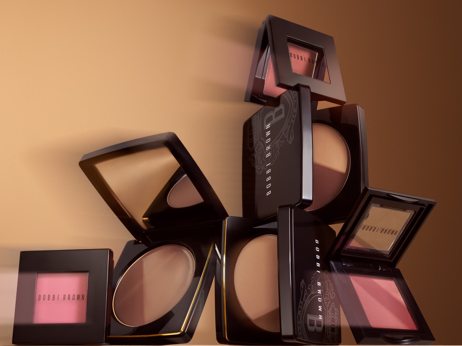 Na beżowym tle ułożone są kwadratowe pudry Bobbi Brown w czarnych opakowaniach, niektóre otwarte, ukazujące odcienie różu i brązu.