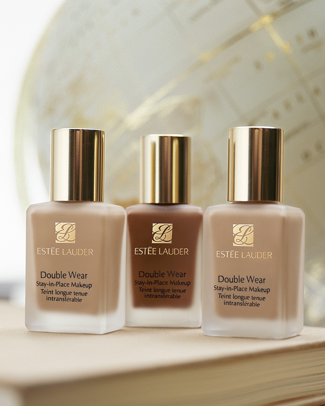Trzy butelki podkładu Estée Lauder Double Wear w odcieniach beżu i brązu, z złotymi nakrętkami, obiecujące długotrwały efekt i komfort noszenia.