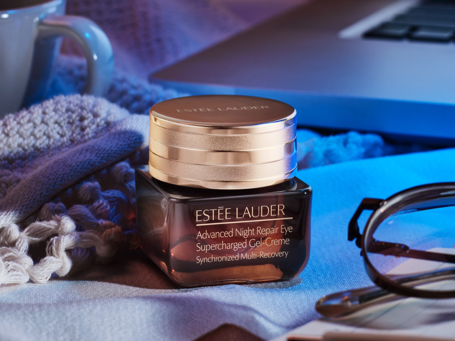 Krem pod oczy Estée Lauder Advanced Night Repair Eye Supercharged Gel-Creme w brązowym słoiczku ze złotą nakrętką, obok leżą okulary, a w tle widać laptopa i kubek.