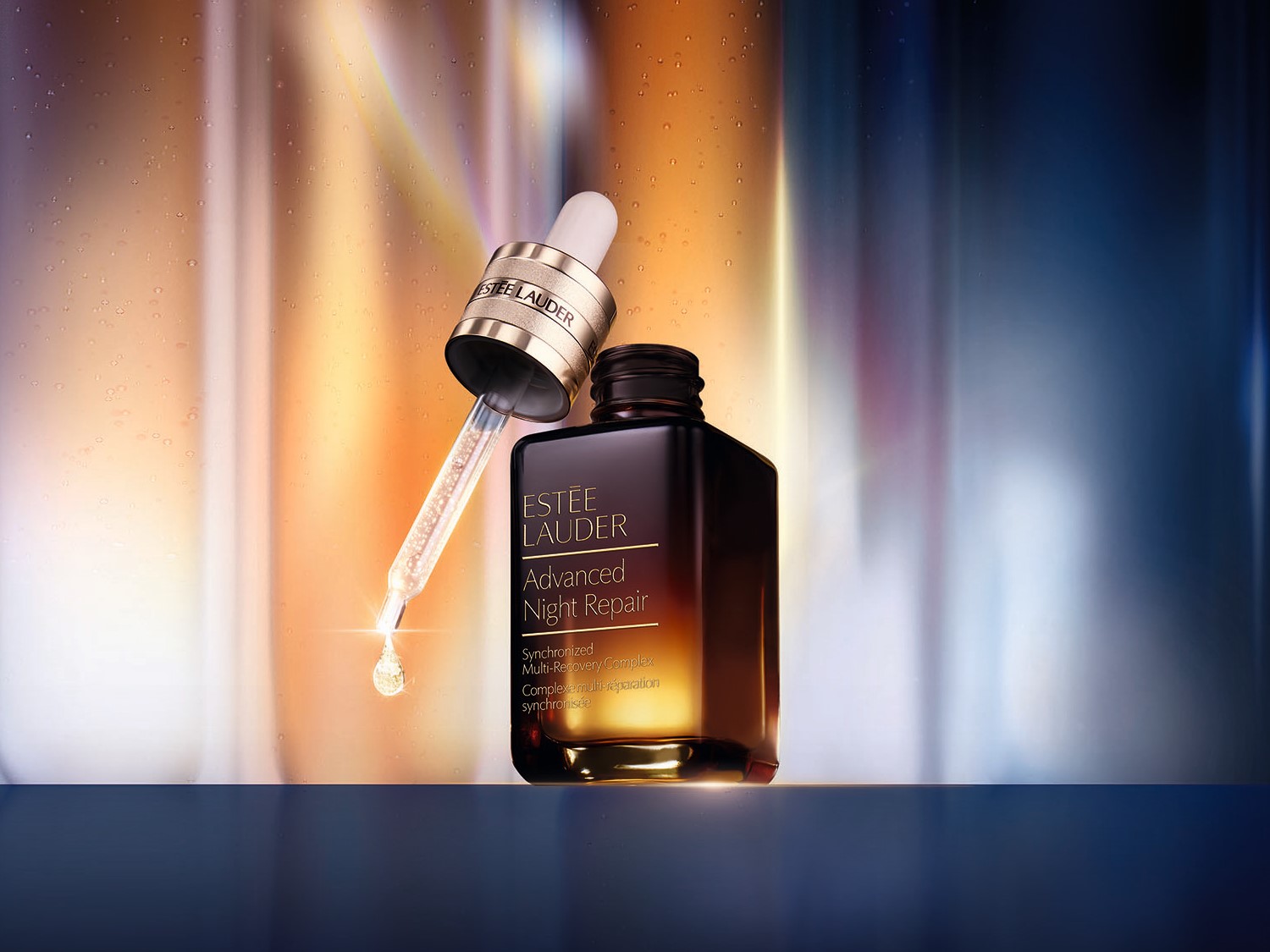Butelka serum Estée Lauder Advanced Night Repair z pipetą, na której wisi kropla produktu, na rozmytym tle w odcieniach złota i błękitu.
