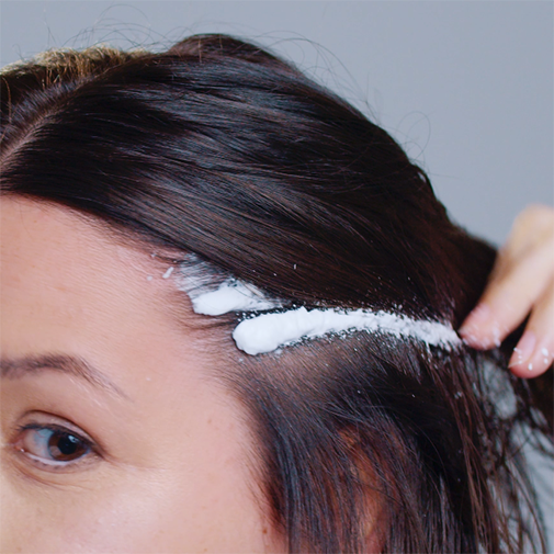 Nahaufnahme von dunklem Haar, auf das eine weiße, schaumige Substanz entlang des Haaransatzes aufgetragen wird.