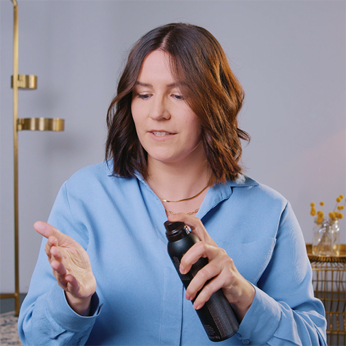 Eine Frau mit gewelltem, braunem Haar sprüht ein Produkt aus einer schwarzen Sprühflasche auf ihre Hand, um die Textur zu zeigen.