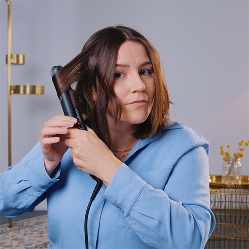Eine Frau mit einem blauen Hemd benutzt ein schwarzes Glätteisen, um ihre braunen, schulterlangen Haare zu stylen.