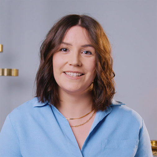 Eine lächelnde Frau mit braunen, gewellten Haaren und einem hellblauen Hemd, die eine goldene Halskette trägt.