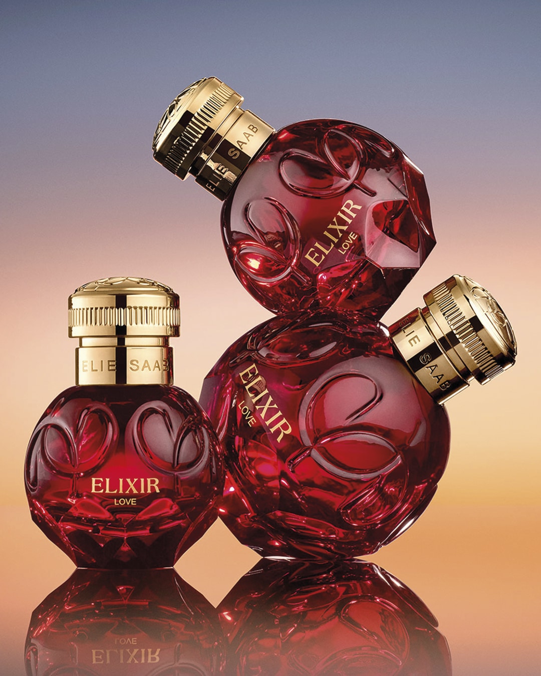 Trzy flakony perfum ELIE SAAB Elixir Love w odcieniach czerwieni i złota, ułożone jeden na drugim, tworzą elegancką kompozycję.
