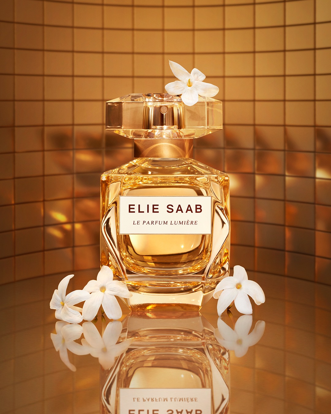 Bursztynowy flakon perfum Elie Saab Le Parfum Lumière, ozdobiony białymi kwiatami, emanuje luksusem i elegancją na złotym tle.