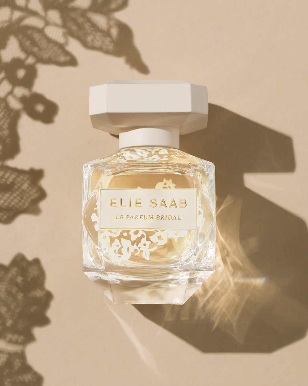Przezroczysty flakon perfum Elie Saab Le Parfum Bridal z beżową etykietą i korkiem, ozdobiony białymi kwiatami, stoi na beżowym tle z cieniem koronki.