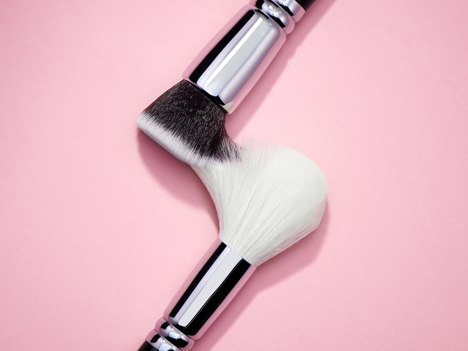 Zwei Make-up-Pinsel mit silbernen Griffen und weichen Borsten liegen auf einem rosa Hintergrund.