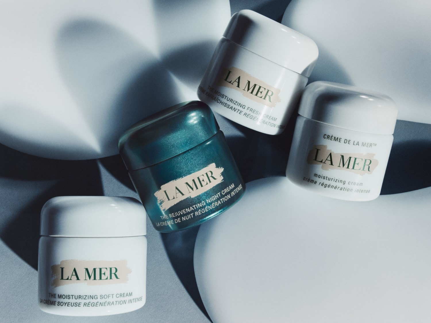 Cztery kremy La Mer w białych i turkusowych opakowaniach, w tym nawilżający krem, świeży krem i krem na noc, obiecujące intensywną regenerację.