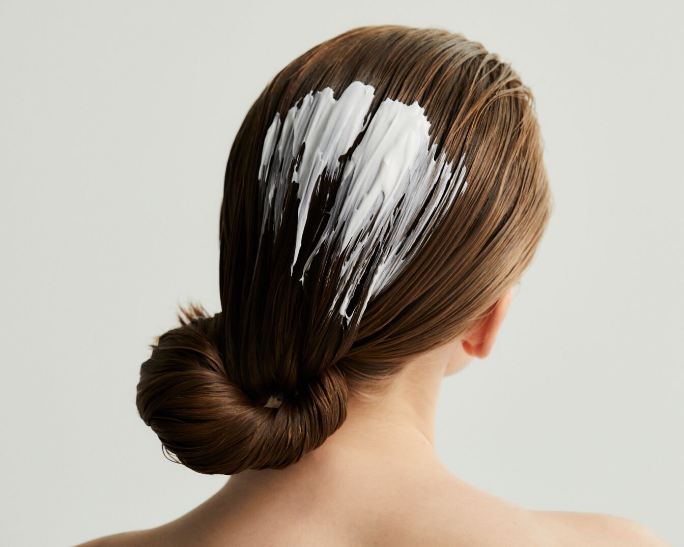 Nahaufnahme der Rückansicht einer Frau mit braunem Haar, auf das eine dicke, weiße Haarmaske aufgetragen wurde, während es zu einem Knoten gezwirbelt ist.