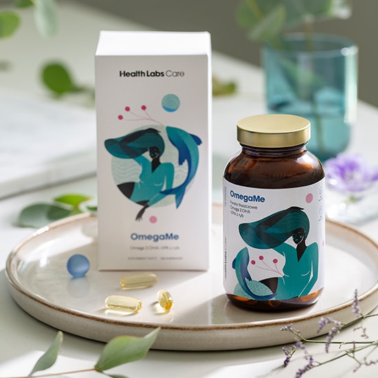 Na ceramicznej tacy stoi brązowa butelka OmegaMe z etykietą przedstawiającą kobietę i rybę, obok niej białe pudełko i kapsułki.