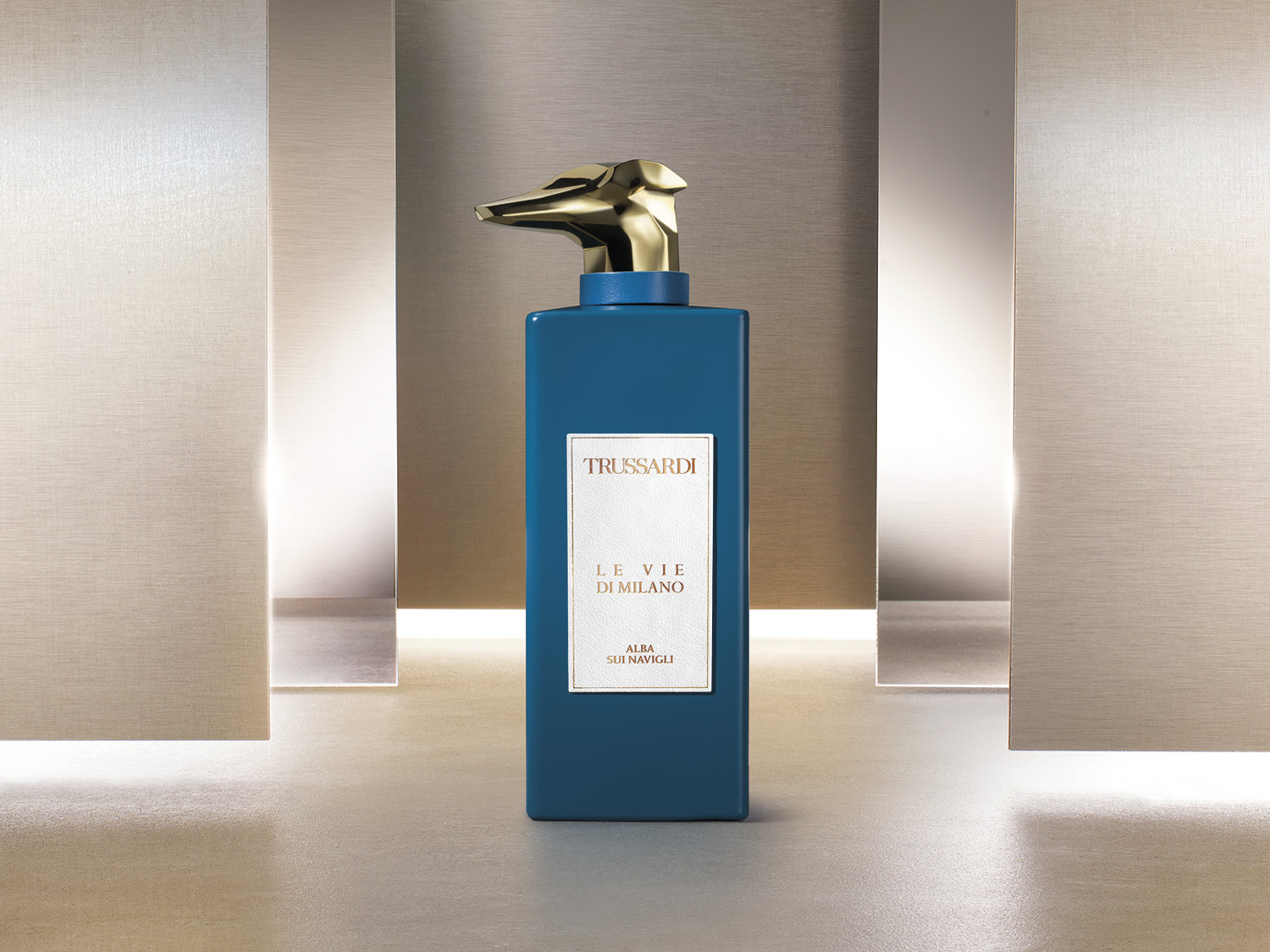 Flakon perfum Trussardi Le Vie Di Milano Alba Sui Navigli ma kształt prostokątnej, ciemnoturkusowej butelki z złotą nakrętką w kształcie głowy charta.