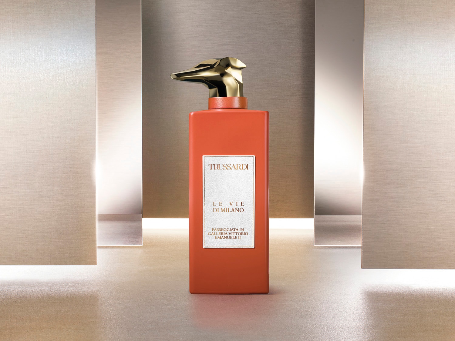 Pomarańczowy flakon perfum Trussardi Le Vie Di Milano z złotą nakrętką w kształcie głowy charta, na etykiecie nazwa perfum.