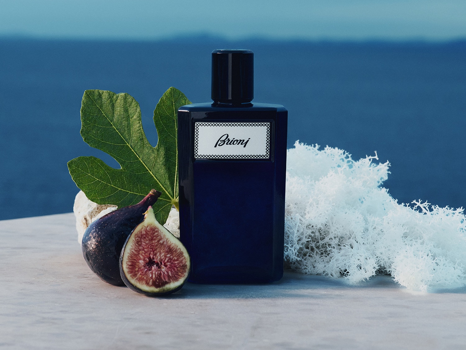 Ciemnoniebieski flakon perfum Brioni z białą etykietą, otoczony świeżymi figami, zielonym liściem figowym i białym mchem, na tle błękitnego morza.