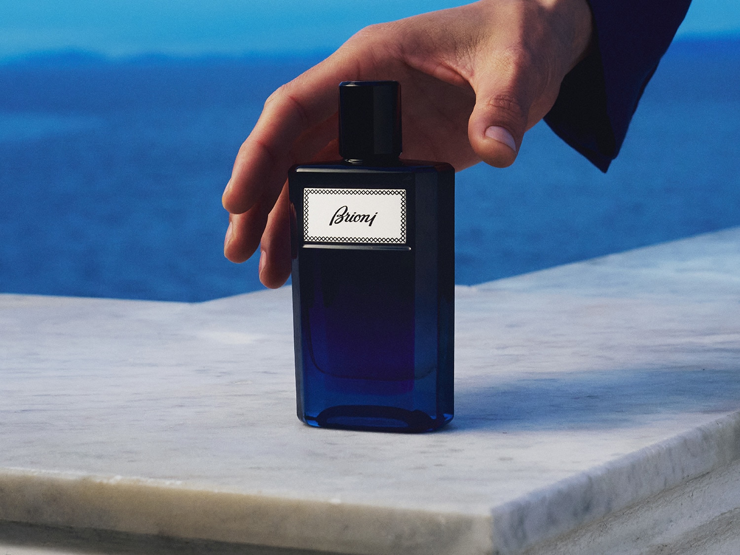 Męska dłoń w eleganckiej koszuli sięga po flakon perfum Brioni w odcieniach granatu, stojący na marmurowej powierzchni, z rozmytym błękitnym tłem przypominającym morze.