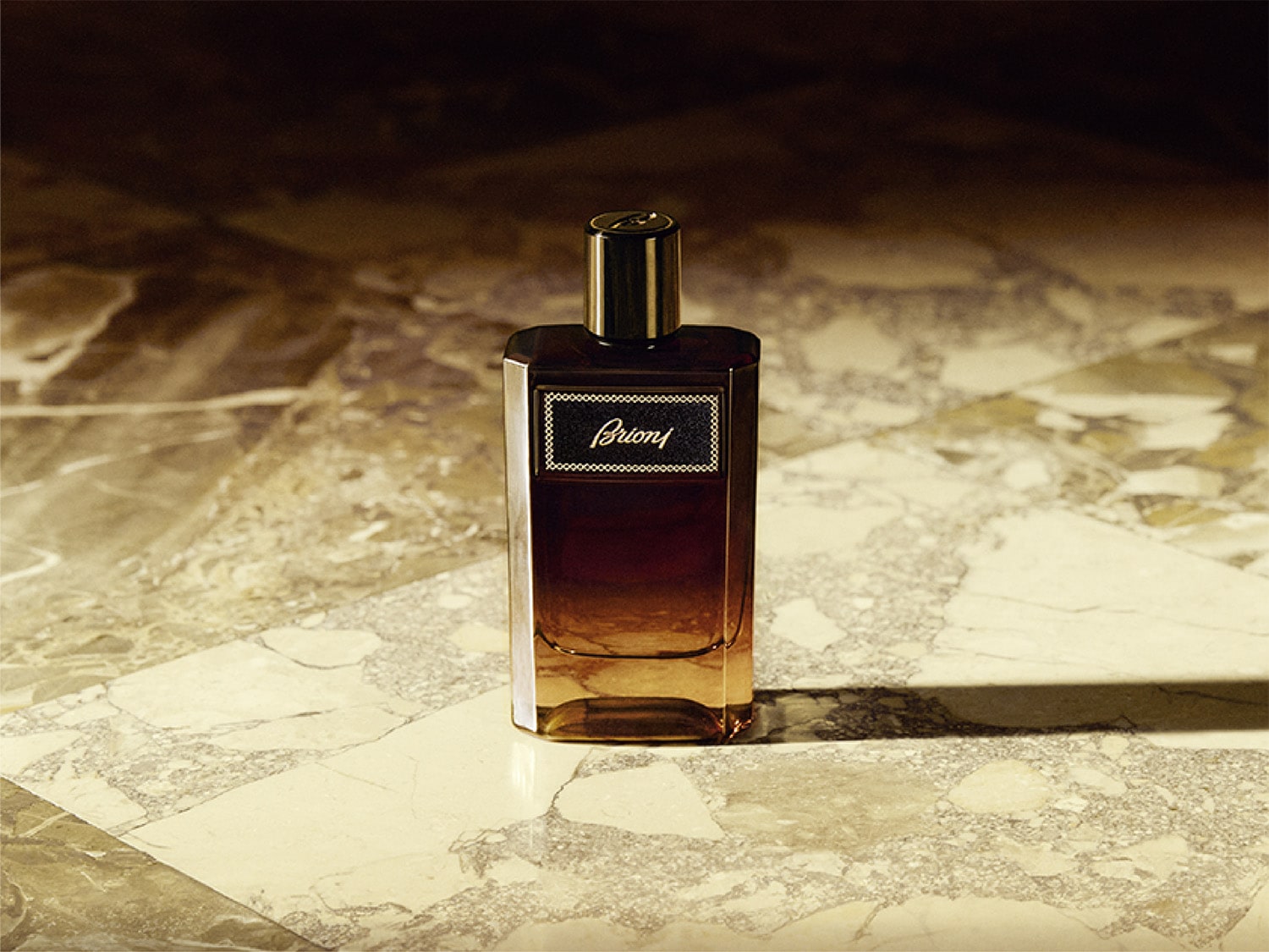 Flakon perfum Brioni w kształcie prostokąta z ciemnobrązowym płynem i złotą nakrętką, stojący na marmurowym blacie.