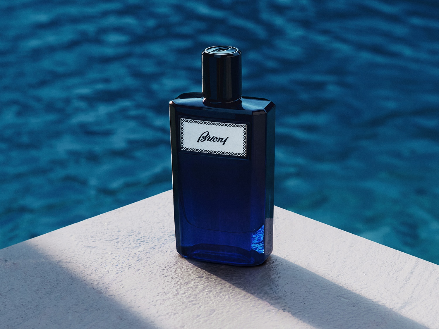 Flakon perfum Brioni w kolorze głębokiego błękitu z czarną nakrętką i białą etykietą z napisem "Brioni" w eleganckiej czcionce, stoi na jasnej powierzchni przy basenie z turkusową wodą.