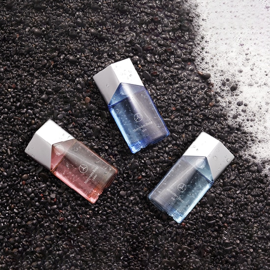 Trzy flakony perfum Mercedes-Benz w odcieniach różu i błękitu, z srebrnymi nakrętkami, leżą na czarnym, kamienistym tle.