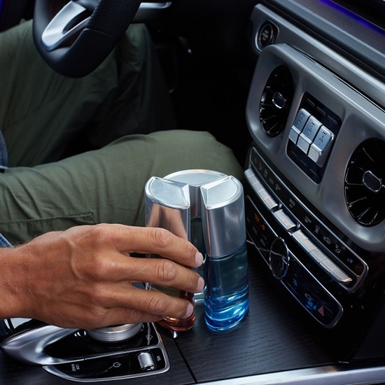 Wnętrze samochodu z dwoma flakonami perfum Mercedes-Benz, jeden w odcieniach pomarańczy, drugi w błękicie, umieszczone w uchwycie na napoje.