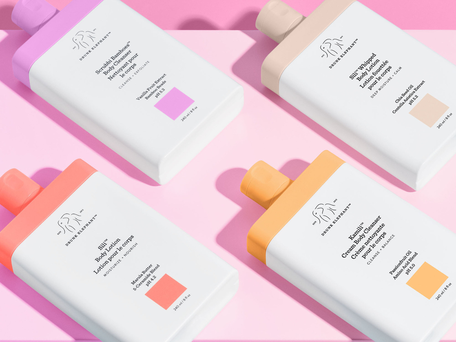Vier witte flessen Drunk Elephant lichaamsreinigers en lotions met kleurrijke accenten op een roze achtergrond, elk met unieke ingrediënten voor reiniging en hydratatie.