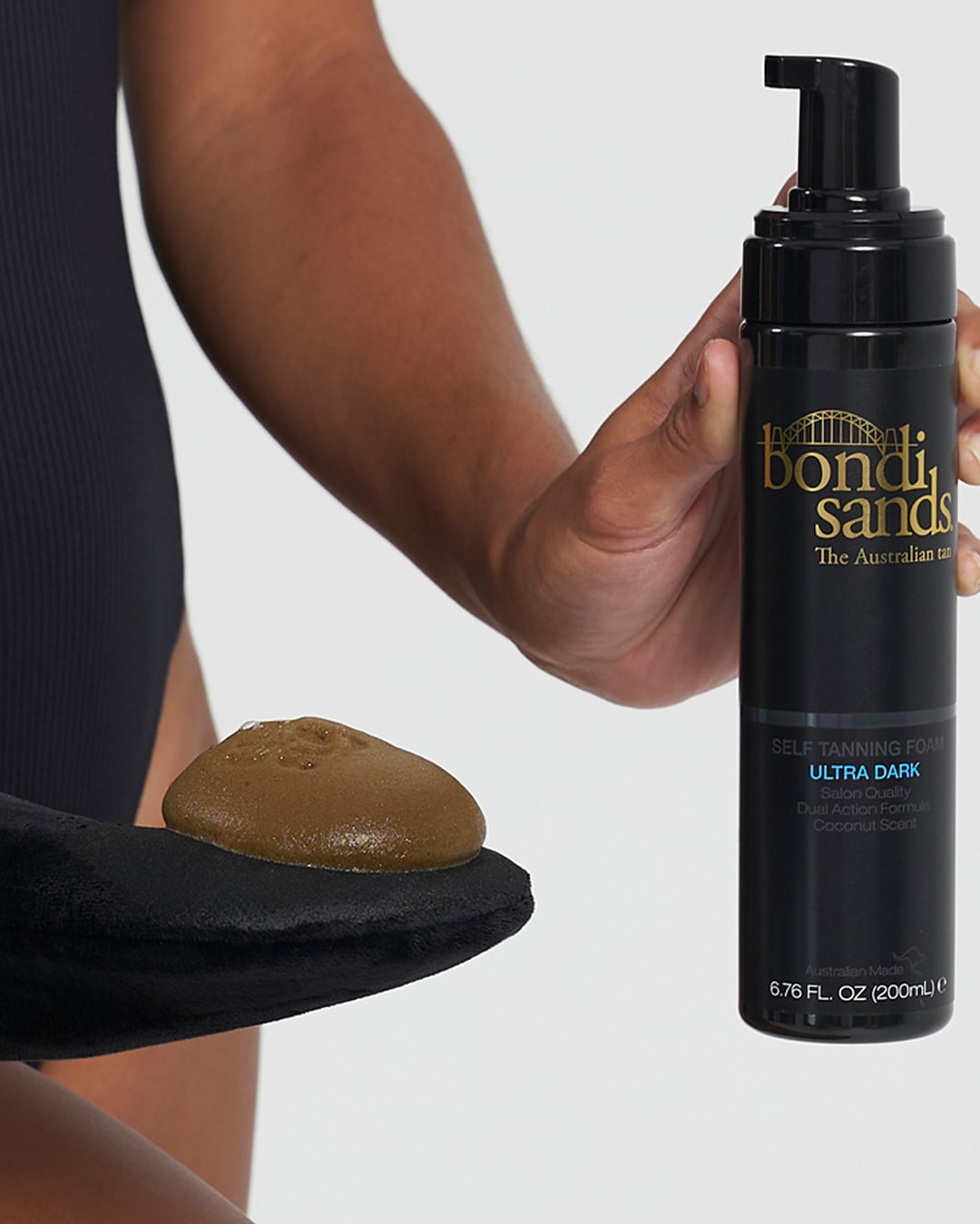 Een donker getinte persoon houdt een zwarte flacon Bondi Sands zelfbruinende mousse vast, terwijl een klodder van de bruine mousse op een zwarte applicator ligt.