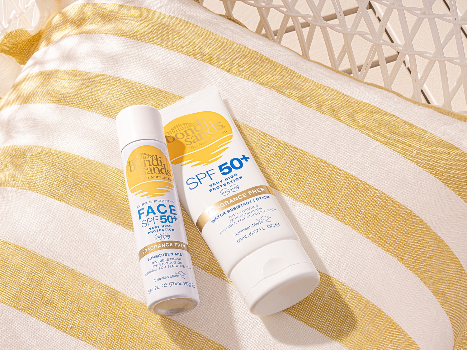 Bondi Sands gezicht SPF 50+ zonnebrandmist en waterbestendige lotion met vitamine E, beide geurvrij en geschikt voor de gevoelige huid, liggen op een geel gestreepte handdoek.