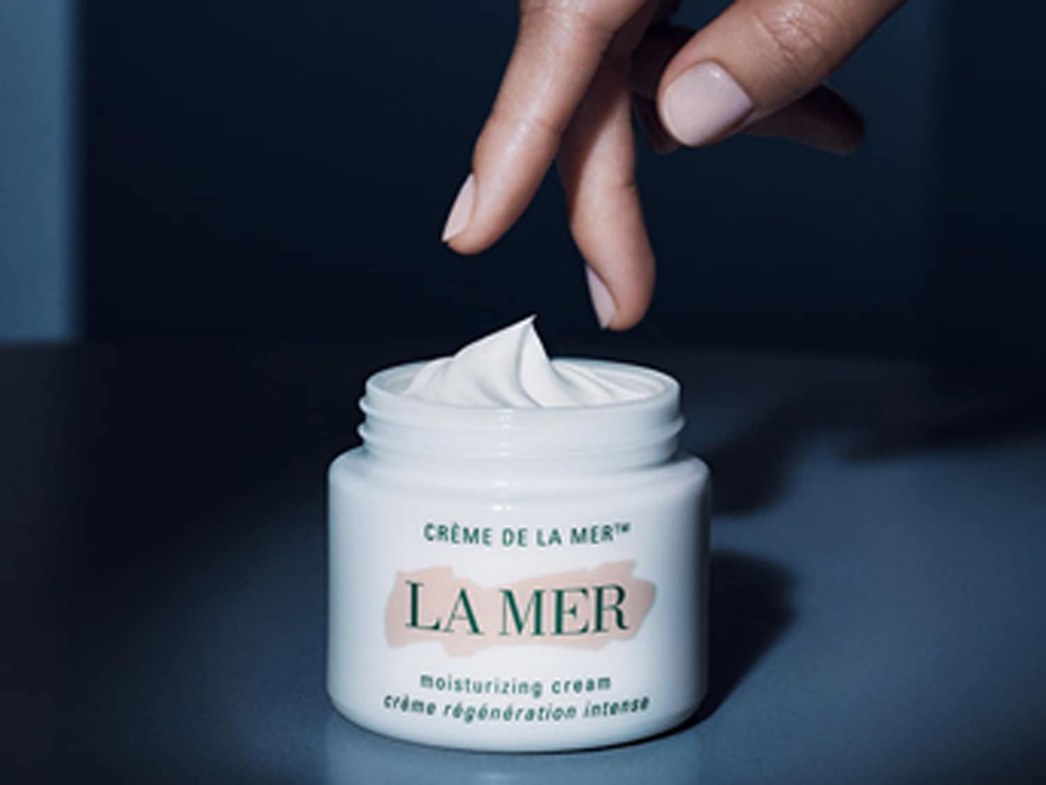 Een hand reikt naar een pot La Mer crème, de witte crème is zichtbaar en klaar voor gebruik, voor een intense hydratatie.