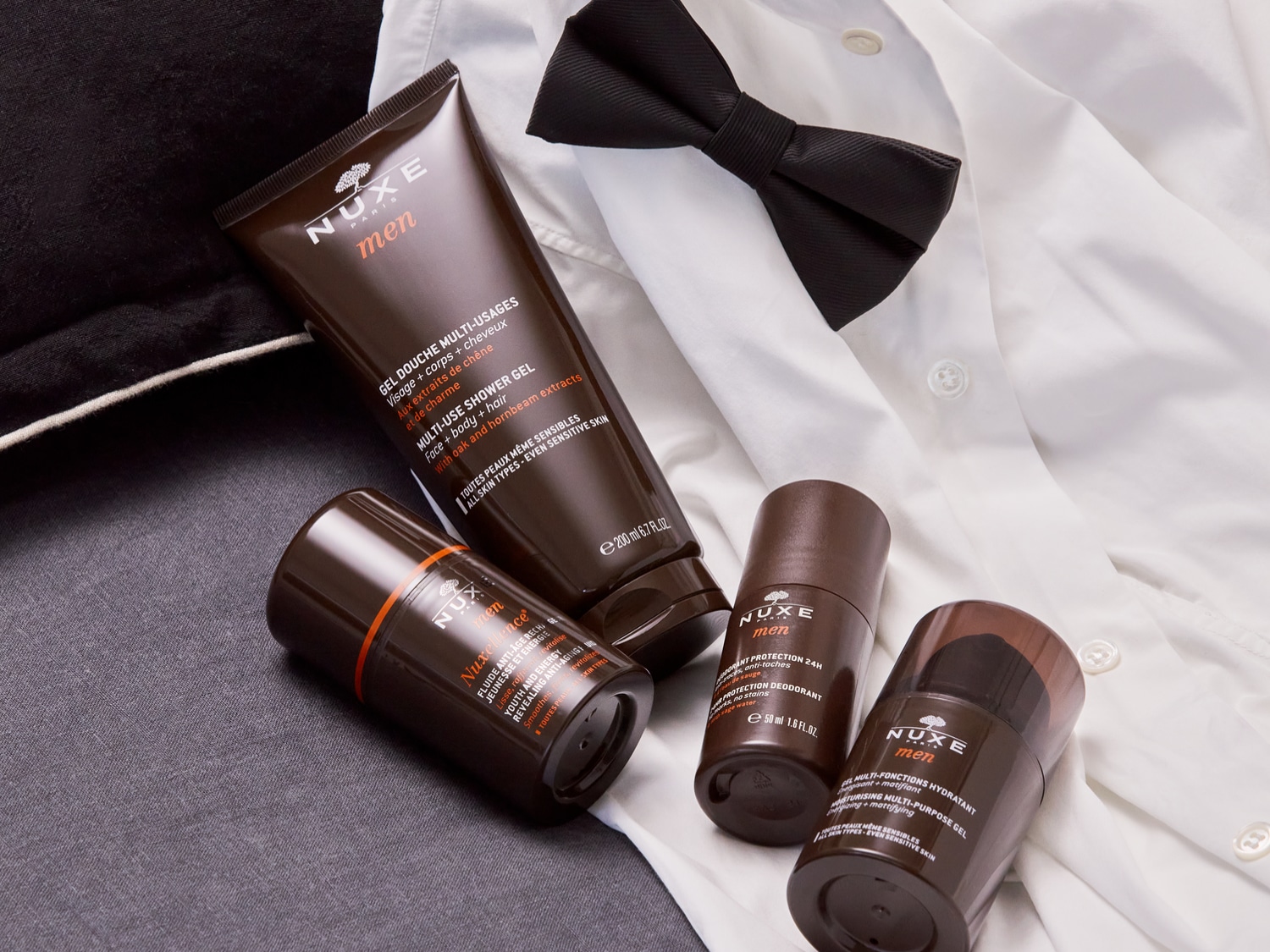 En la imagen se aprecia una colección de productos Nuxe Men sobre una camisa blanca y un elegante corbatín negro, incluyendo gel de ducha multiuso y desodorantes, resaltando el cuidado personal masculino con un toque de sofisticación.