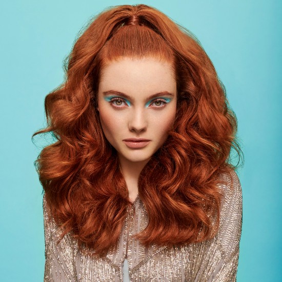 Na turkusowym tle kobieta z rudymi, kręconymi włosami i niebieskim makijażem oczu, ubrana w błyszczącą bluzkę, prezentuje modny wygląd.