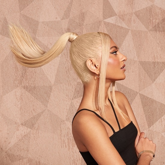 Profil kobiety z blond włosami upiętymi w wysoki kucyk, z delikatnym makijażem i czarną bluzką na ramiączkach, na tle w odcieniach beżu.