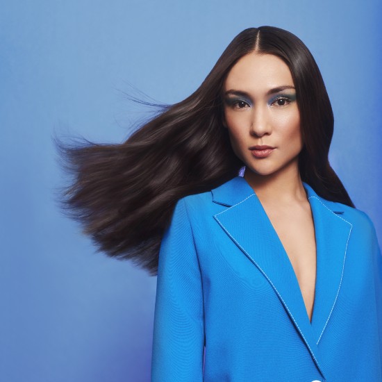 Kobieta z długimi, ciemnymi włosami i niebieskim makijażem oczu, ubrana w niebieską marynarkę, pozuje na niebieskim tle.