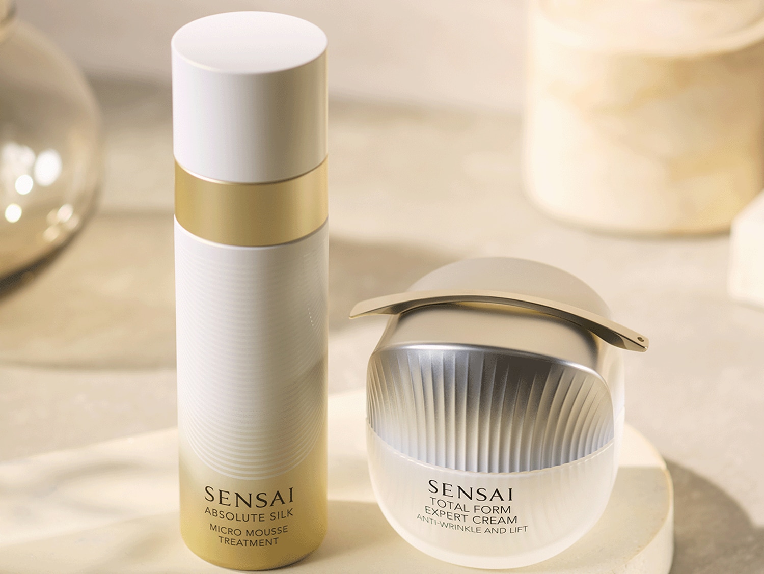 Dwa produkty Sensai, Absolute Silk Micro Mousse Treatment w białej butelce ze złotym paskiem i Total Form Expert Cream w eleganckim, srebrno-białym słoiczku, stoją na beżowym tle, gotowe do pielęgnacji przeciwzmarszczkowej i liftingującej.